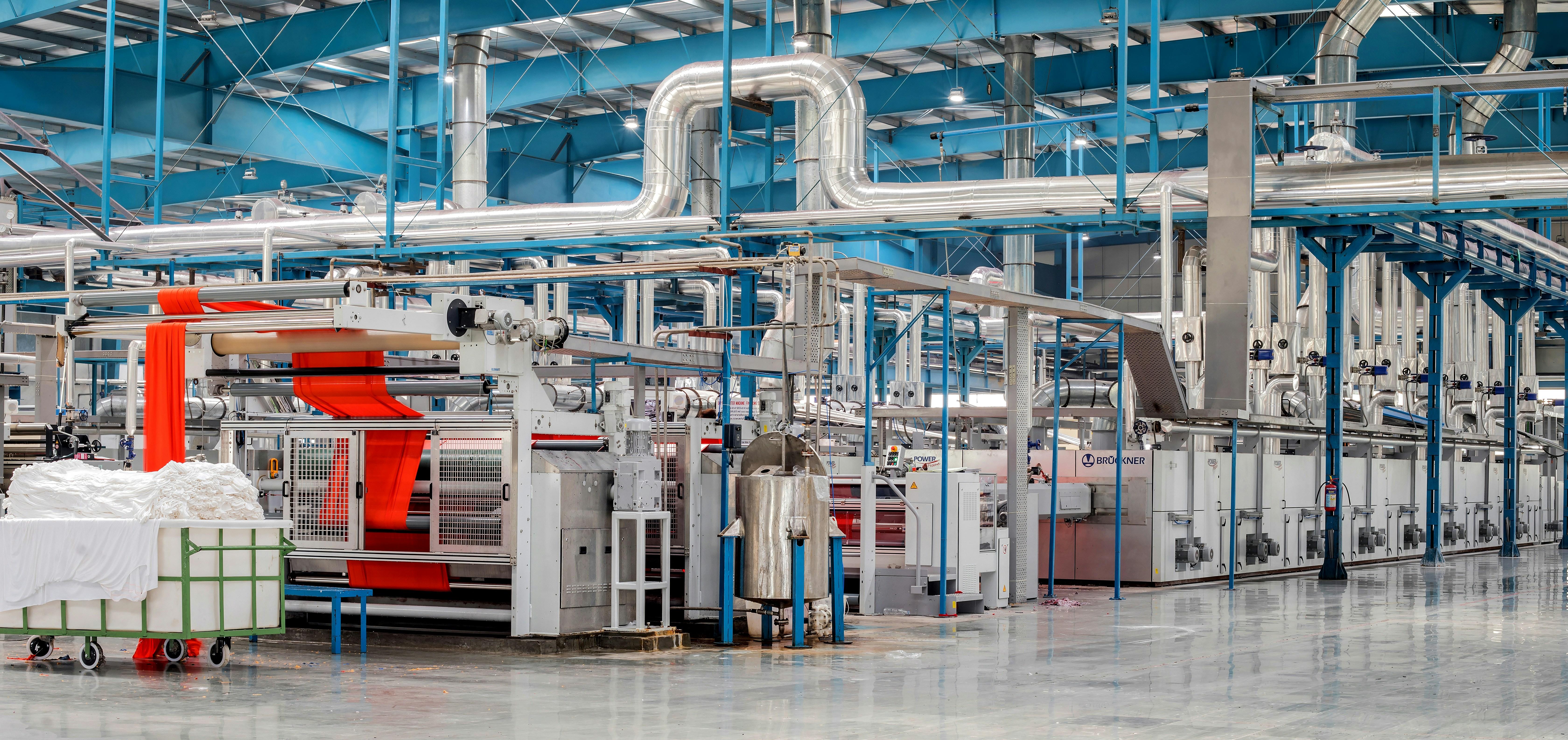 Textile factory floor with fabric-finishing machinery, a run of red cloth feeding through a roller line