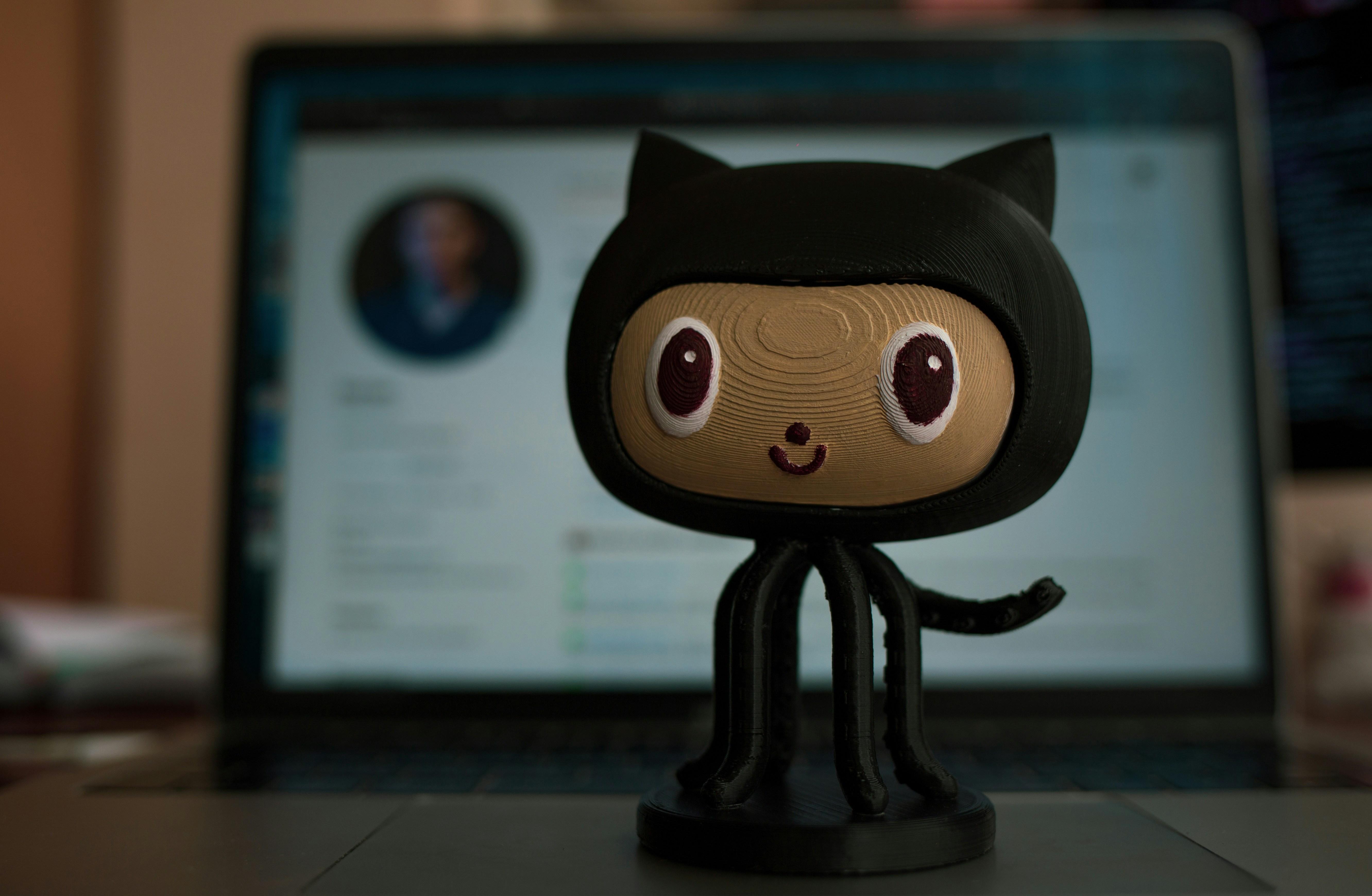 GitHub Octocat figurine standing in front of a laptop showing a GitHub profile